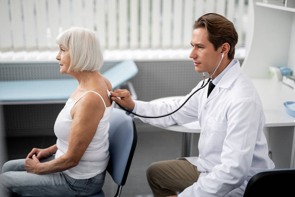 Calm aged woman sitting on the chair and professional general practitioner using stethoscope during the examination - Foto, Imagen