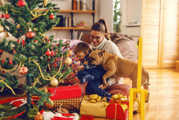 Mother and son playing with dog while sitting on the floor. Next to them Christmas tree and gifts. Home interior, Christmas holidays concept. - Photo, image