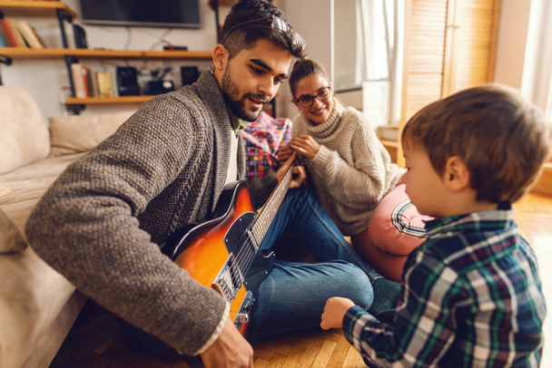 Padre tocando la guitarra eléctrica y sentado en el suelo mientras su familia lo mira. Interior del hogar
. - Foto, imagen