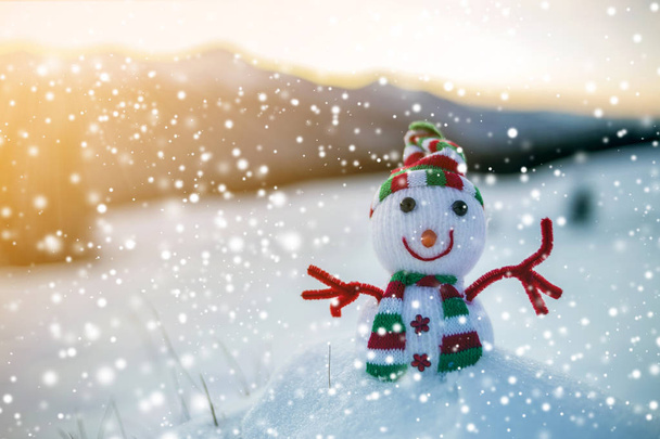 Pequeño juguete divertido muñeco de nieve bebé en sombrero de punto y bufanda en nieve profunda al aire libre en las montañas borrosas paisaje y la caída de grandes copos de nieve de fondo. Feliz Año Nuevo y Feliz Navidad tarjeta de felicitación tema. - Foto, imagen