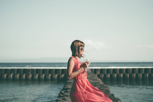 Genç kadın uzun kırmızı bir elbise ve bir kadeh şarap deniz arka plan üzerinde poz ile. - Fotoğraf, Görsel