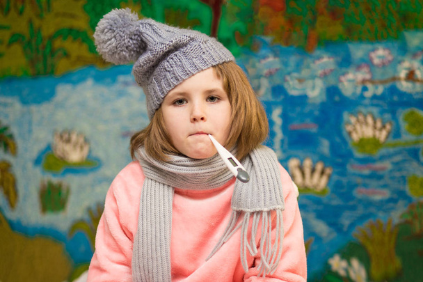 Άρρωστο κοριτσάκι με θλιμμένο πρόσωπο και το θερμόμετρο στο στόμα φθορά σε κασκόλ και καπέλο - Φωτογραφία, εικόνα