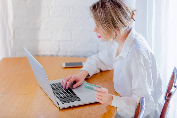 Mladá dívka v bílé košili, sedí v tabulce s přenosným počítačem a držení platební karty. Podnikání, vzdělávání, nakupování a další koncepce - Fotografie, Obrázek