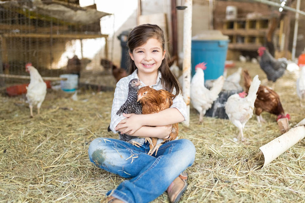 Pleine longueur de petite fille souriante tenant des poulets tout en étant assis sur le foin à la ferme avicole
 - Photo, image
