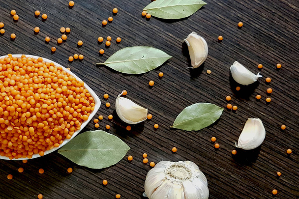 Red lentils in a white bowl on a dark wooden background, next to garlic and leaves on the laurel overlooking the top view. - Foto, immagini