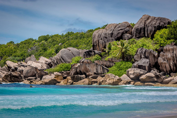 Increíble escenario de playa con mar de agua azul y roca de forma única entre la vegetación verde en Praslin en Seychelles en un día de cielo azul
 - Foto, Imagen