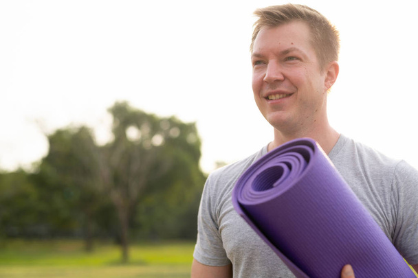 Young happy thoughtful man with yoga mat at the park - Foto, Imagen