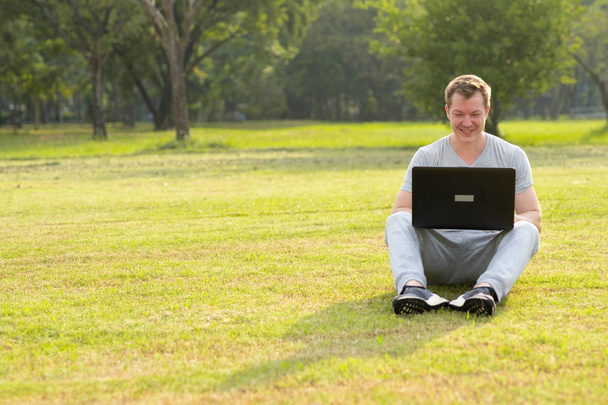Junger glücklicher Mann sitzt mit Laptop im Park - Foto, Bild