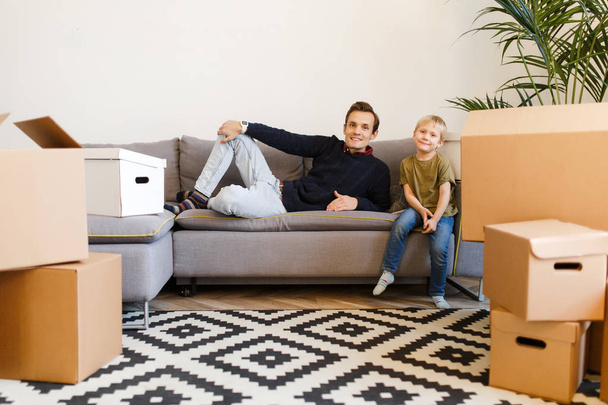 Photo of happy father and son sitting on gray sofa among cardboard boxes - Photo, Image