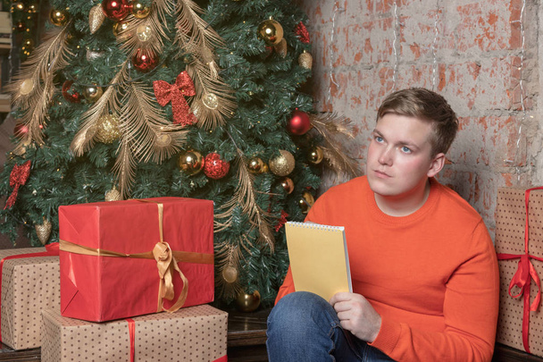 Handsome guy is writing a letter to Santa sitting under the tree surrounded by boxes of gifts - Fotoğraf, Görsel