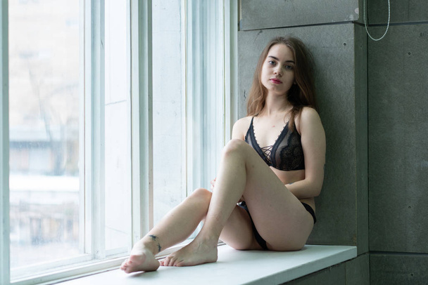 nuori kaunis tyttö poseeraa huoneessa, istuu huoneen lähellä ikkunan musta alusvaatteet
 - Valokuva, kuva