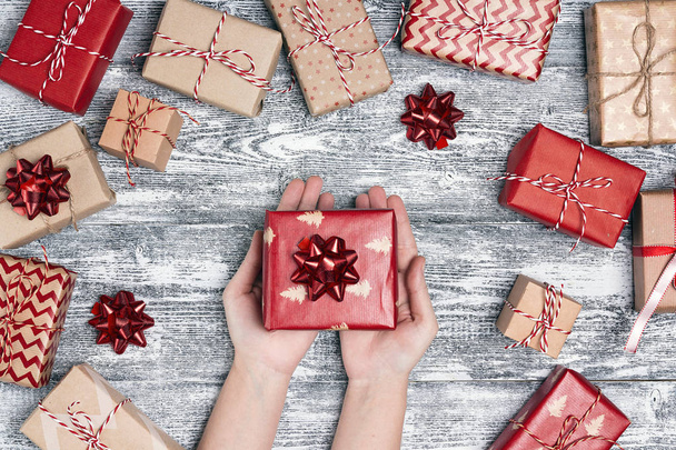 Women's hands with gift box surrounded by wrapped gifts. Top-down Christmas composition on grey wooden background. - 写真・画像