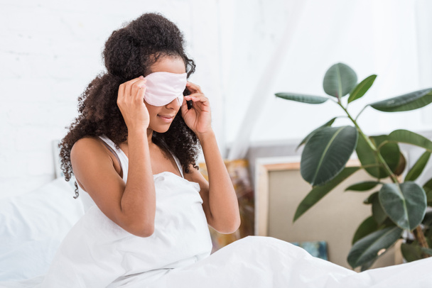 young african american girl with eyes covered by sleeping blindfold in bed during morning time at home - Foto, afbeelding