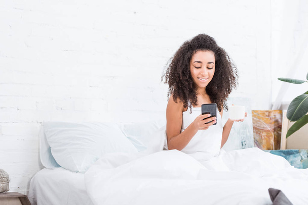 巻き毛の少女を浮かべて朝時に自宅のベッドでスマート フォンを使用してコーヒー カップ - 写真・画像