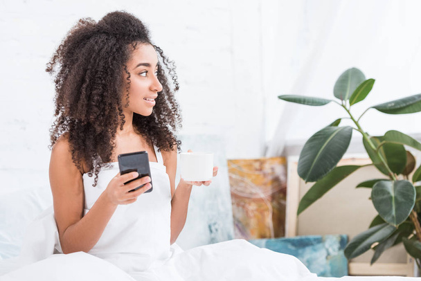 africká americká mladá žena s pomocí smartphone v posteli doma v době Ranní šálek kávy - Fotografie, Obrázek