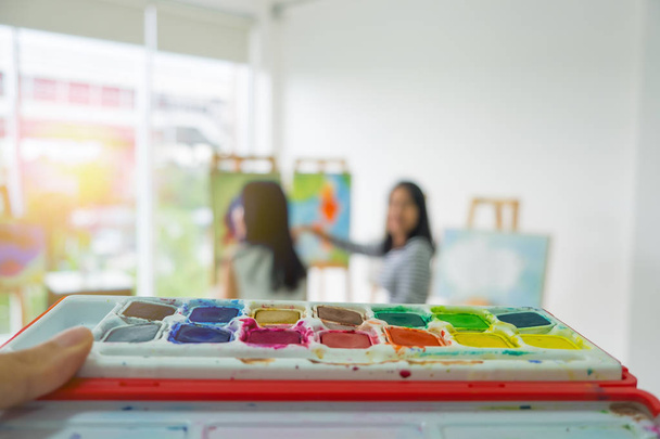 close up hand holding a palette for art. With a background blur two young girls are sitting drawing. copy space. - 写真・画像