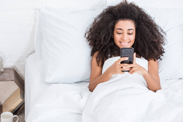 joyful african american young woman using smartphone in bed during morning time at home - Photo, Image