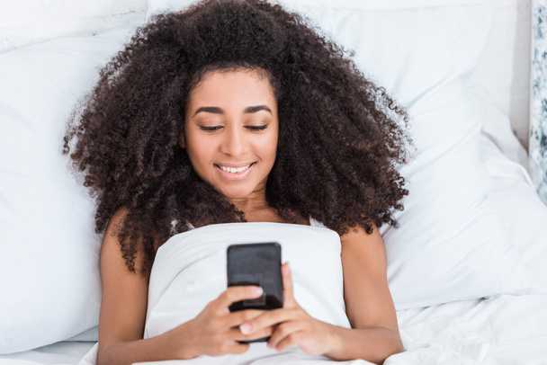 boldog afrikai-amerikai fiatal nő használ smartphone az ágyban reggel időben otthon - Fotó, kép