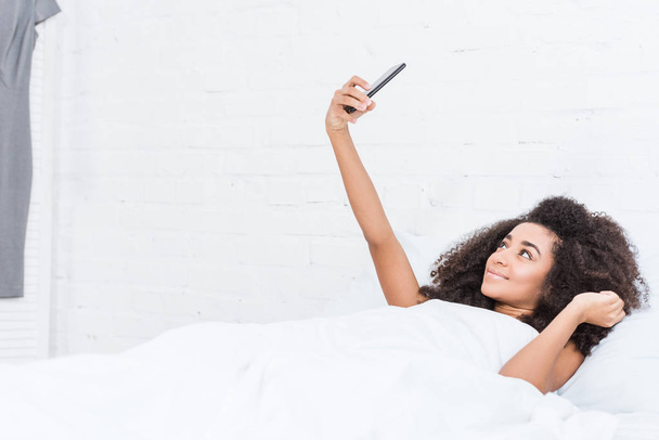 vista lateral da menina americana africana feliz tomando selfie no smartphone na cama durante o tempo da manhã em casa
 - Foto, Imagem