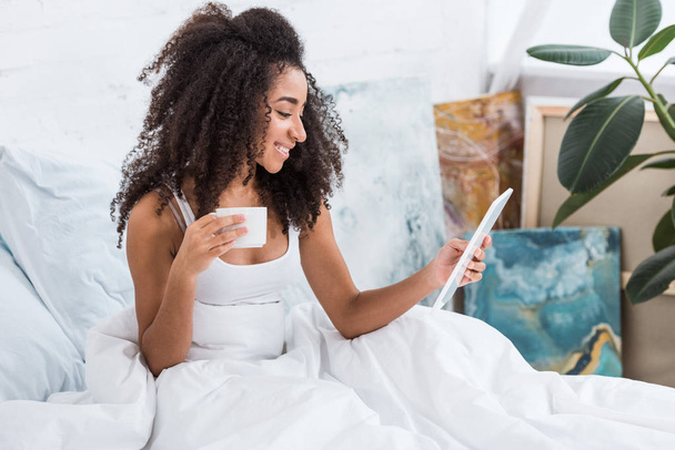 usmívající se africká americká žena pomocí digitálních tabletu a pití kávy v posteli doma - Fotografie, Obrázek