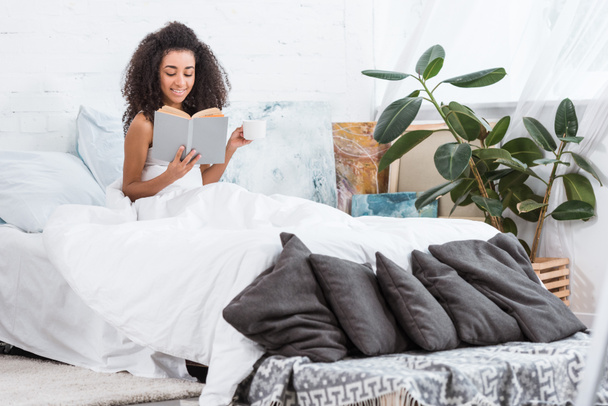 curly african american girl holding coffee cup and reading book in bed during morning time at home - Foto, Imagem