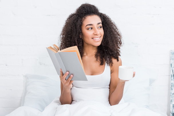 chica afroamericana feliz sosteniendo taza de café y libro de lectura en la cama durante la mañana en casa
 - Foto, Imagen