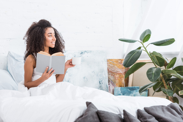 vista laterale della ragazza afro-americana in possesso di tazza di caffè e libro di lettura a letto durante il mattino a casa
 - Foto, immagini