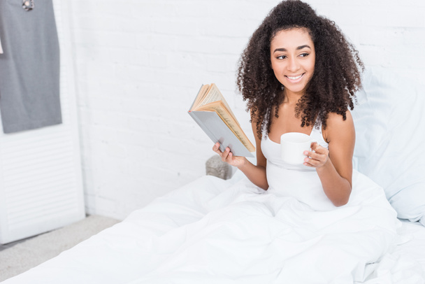 selective focus of african american girl holding coffee cup and reading book in bed during morning time at home - Fotografie, Obrázek