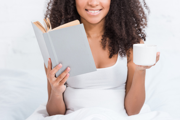 partial view of african american girl holding coffee cup and reading book in bed during morning time at home - Фото, зображення