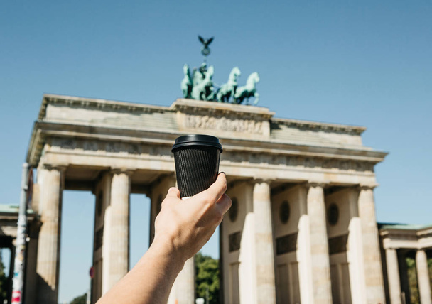 eine person hält eine einwegbecher mit kaffee oder einem anderen getränk auf dem hintergrund des brandenburger tores in berlin. - Foto, Bild