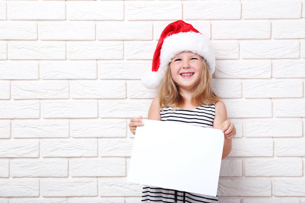 Little girl in santa hat holding blank sheet of paper on brick wall background - Foto, Bild