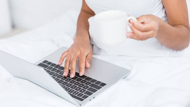 cropped image of woman with coffee cup using laptop in bed during morning time at home - Фото, изображение