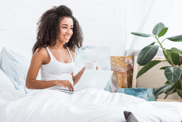 νεαρή γυναίκα κρατώντας φλιτζάνι καφέ και χρησιμοποιώντας το laptop στο κρεβάτι κατά τη διάρκεια του χρόνου το πρωί στο σπίτι - Φωτογραφία, εικόνα