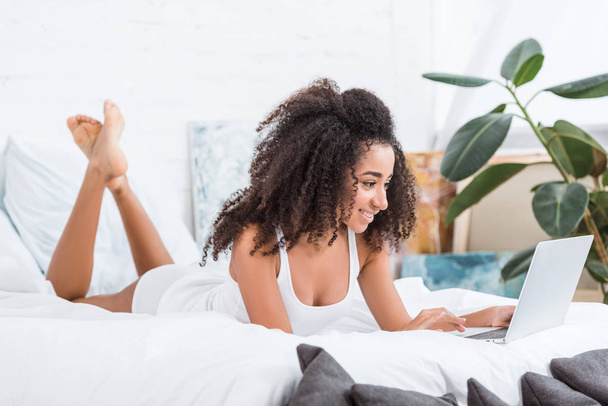 χαρούμενο σγουρά κορίτσι που χρησιμοποιούν φορητό υπολογιστή στο κρεβάτι κατά τη διάρκεια του χρόνου το πρωί στο σπίτι - Φωτογραφία, εικόνα