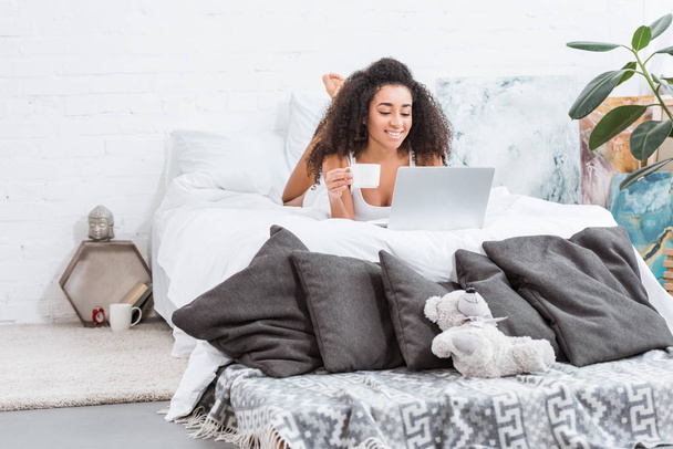 atraktivní šťastná dívka držící šálek kávy a pomocí přenosného počítače v posteli během ranní čas doma - Fotografie, Obrázek