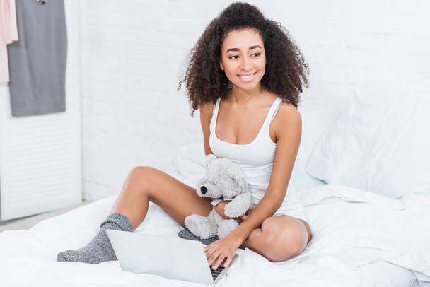 curly young woman holding teddy bear and using laptop in bed during morning time at home - Zdjęcie, obraz