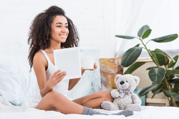 atrakcyjna młoda kobieta Afryki amerykański holding filiżanki kawy i za pomocą cyfrowego tabletu w łóżku z misiem w domu - Zdjęcie, obraz