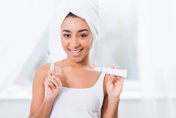 χαρούμενη αφρικανική αμερικανική γυναίκα με πετσέτα τυλιγμένη πάνω από το κεφάλι δείχνει οδοντόκρεμα και οδοντόβουρτσα  - Φωτογραφία, εικόνα