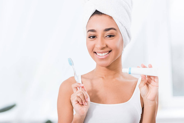 positive african american young woman with towel wrapped over head showing toothpaste and toothbrush  - Photo, Image