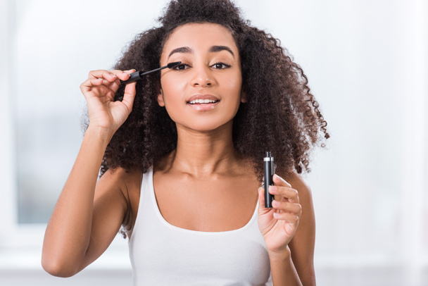 curly african american girl using mascara and looking at camera - Photo, Image