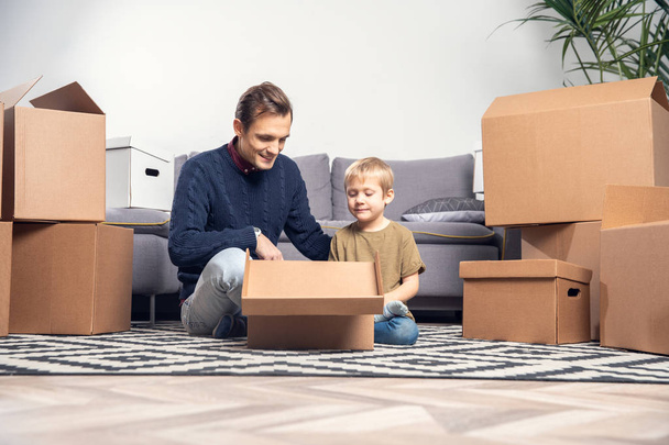 Portrait of man and boy sitting on floor among cardboard boxes in room - Фото, зображення