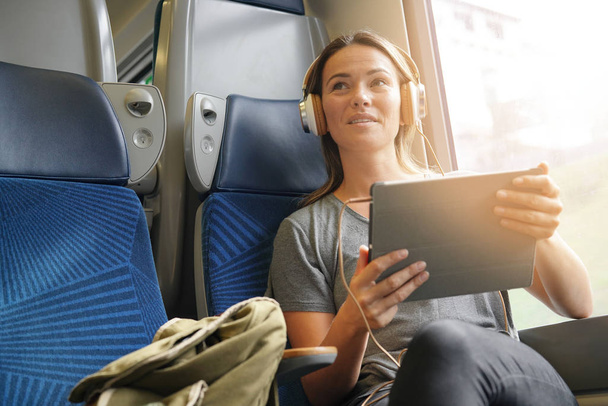 彼女のタブレットとヘッドフォンを電車で旅行する若い女性                               - 写真・画像