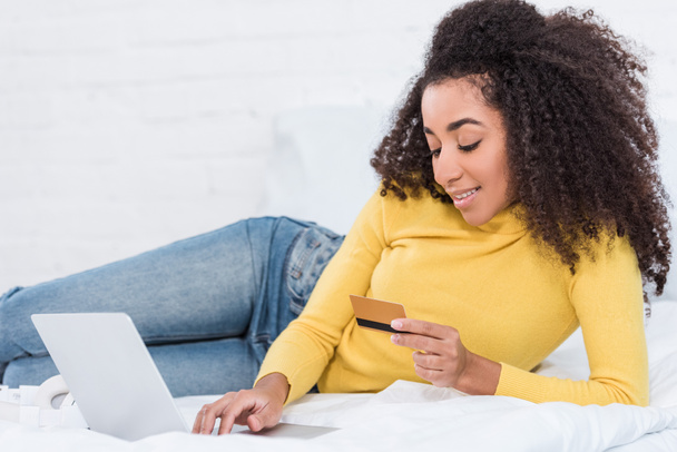 young attractive african american woman holding credit card and doing online shopping on laptop in bed at home - Photo, Image