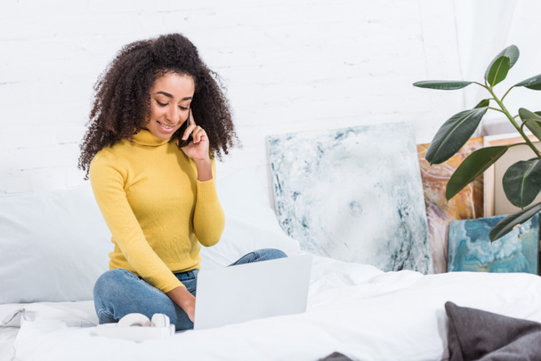 göndör afro-amerikai női szabadúszó smartphone beszélő, és dolgozik a laptop az ágyban otthon - Fotó, kép