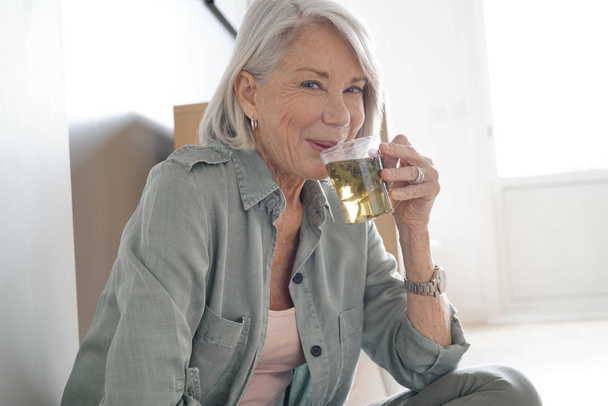  Attractive senior woman sitting on the floor at home with tea                              - Foto, imagen