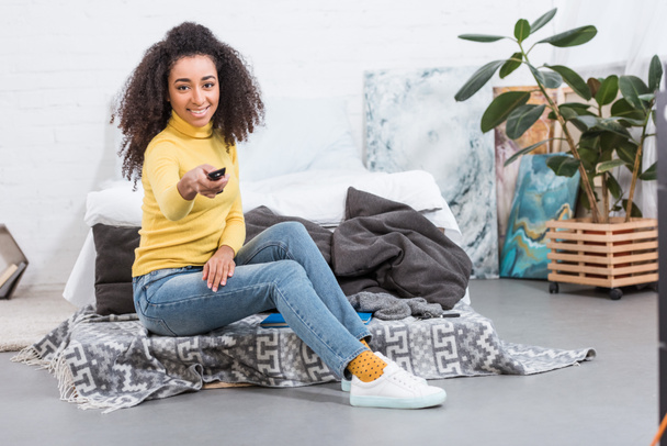 vrolijke stijlvolle Afrikaans Amerikaans meisje met behulp van de afstandsbediening en tv kijken thuis - Foto, afbeelding