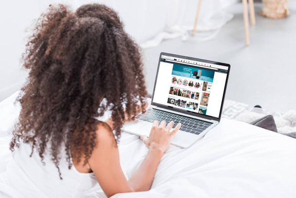 rear view of curly woman using laptop with amazon on screen in bed at home - Zdjęcie, obraz
