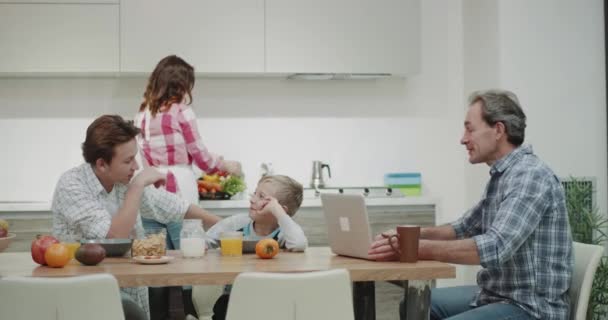 Happy family with mature parents spending a nice breakfast together while mom cooking dad showing a nice video from laptop to all family they all looking through the laptop. 4k - Séquence, vidéo