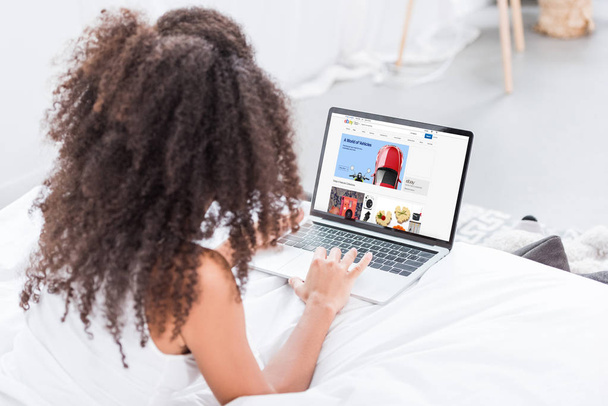 rear view of curly woman using laptop with ebay on screen in bed at home - Zdjęcie, obraz