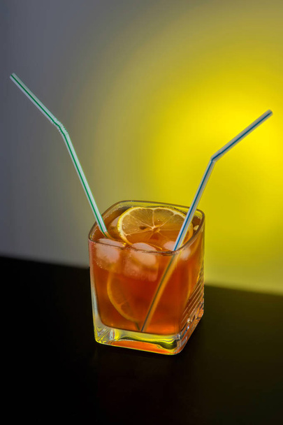Transparent glass of whiskey with lemon, ice and two cocktail straws, close-up against the background of a yellow light source - Zdjęcie, obraz